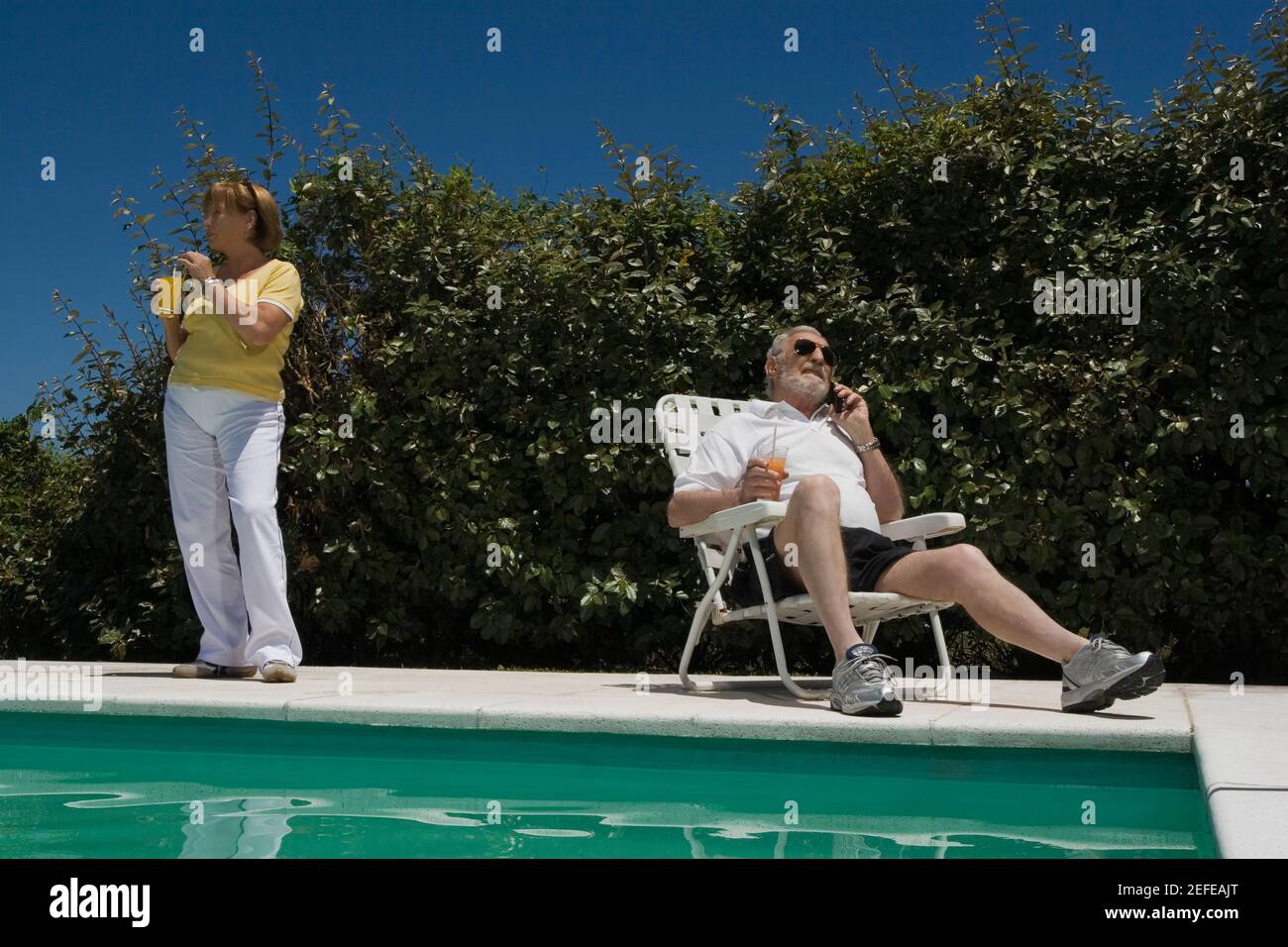 Homme senior parlant sur un téléphone portable et femme senior boire du jus au bord de la piscine Banque D'Images