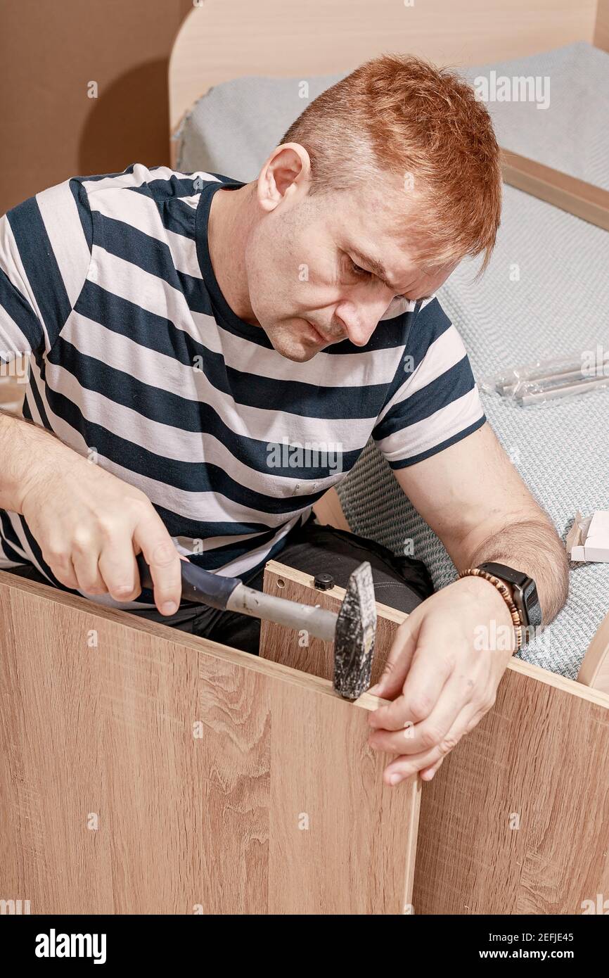 Bon aspect concentré homme caucasien en t-shirt rayé assemble des meubles avec un marteau. Assemblage, réparation, déplacement de meubles. Tir vertical Banque D'Images