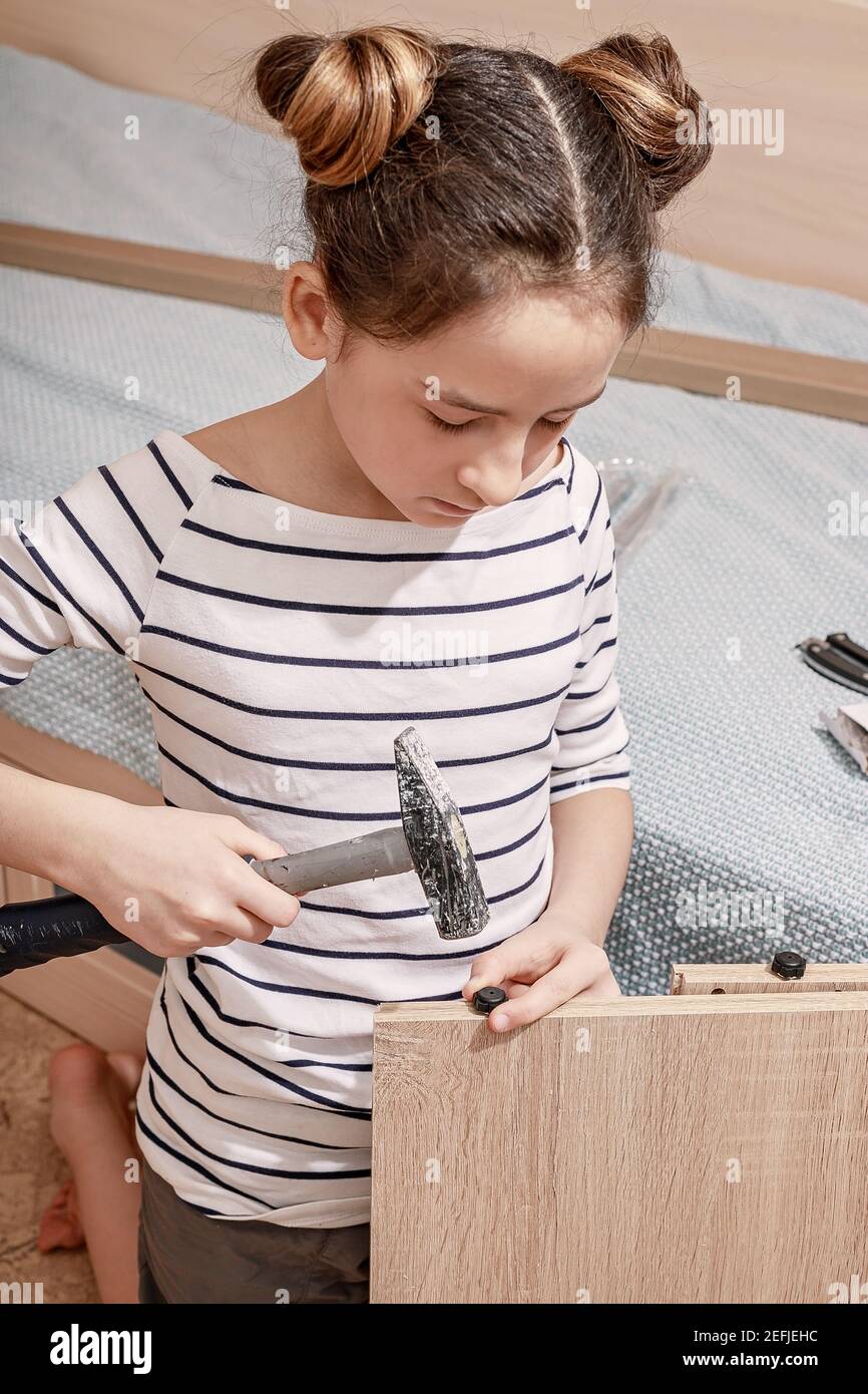 Petite belle fille caucasienne dans une chemise rayée apprend à assembler des meubles avec un marteau. Travaux ménagers. Tir vertical Banque D'Images