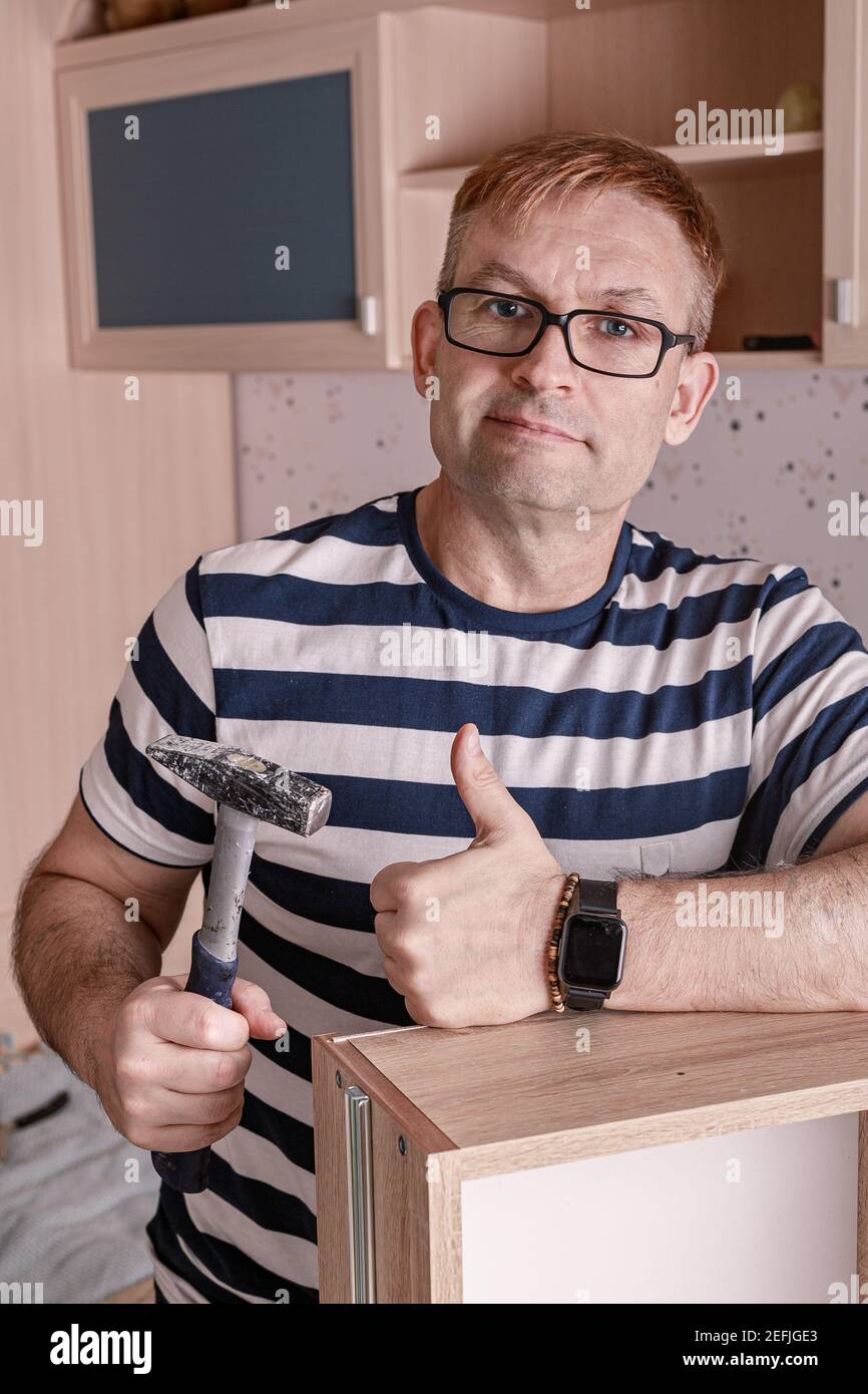Souriant homme caucasien en t-shirt rayé et verres assemble de nouveaux meubles avec un marteau. Homme faisant des travaux ménagers. Tir vertical Banque D'Images