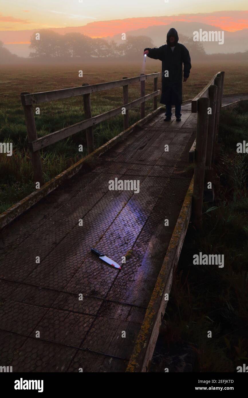 Homme habillé de capot noir tenant la torche et l'éclairage sanglant couteau sur sentier en bois au lever du soleil Banque D'Images