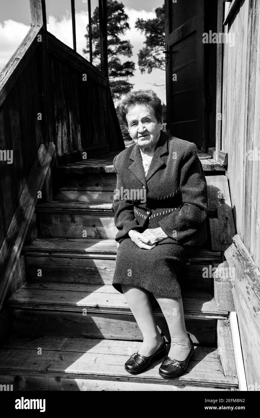 Vieille femme assise sur le porche de sa maison de village. Photo en noir et blanc. Banque D'Images