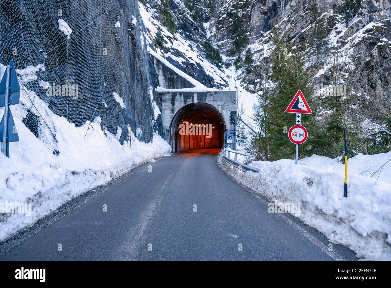 Entrée d'un tunnel éclairé le long d'une route alpine enneigée en hiver. Un panneau d'avertissement contre les chutes de pierres sur le côté droit de la route. Banque D'Images