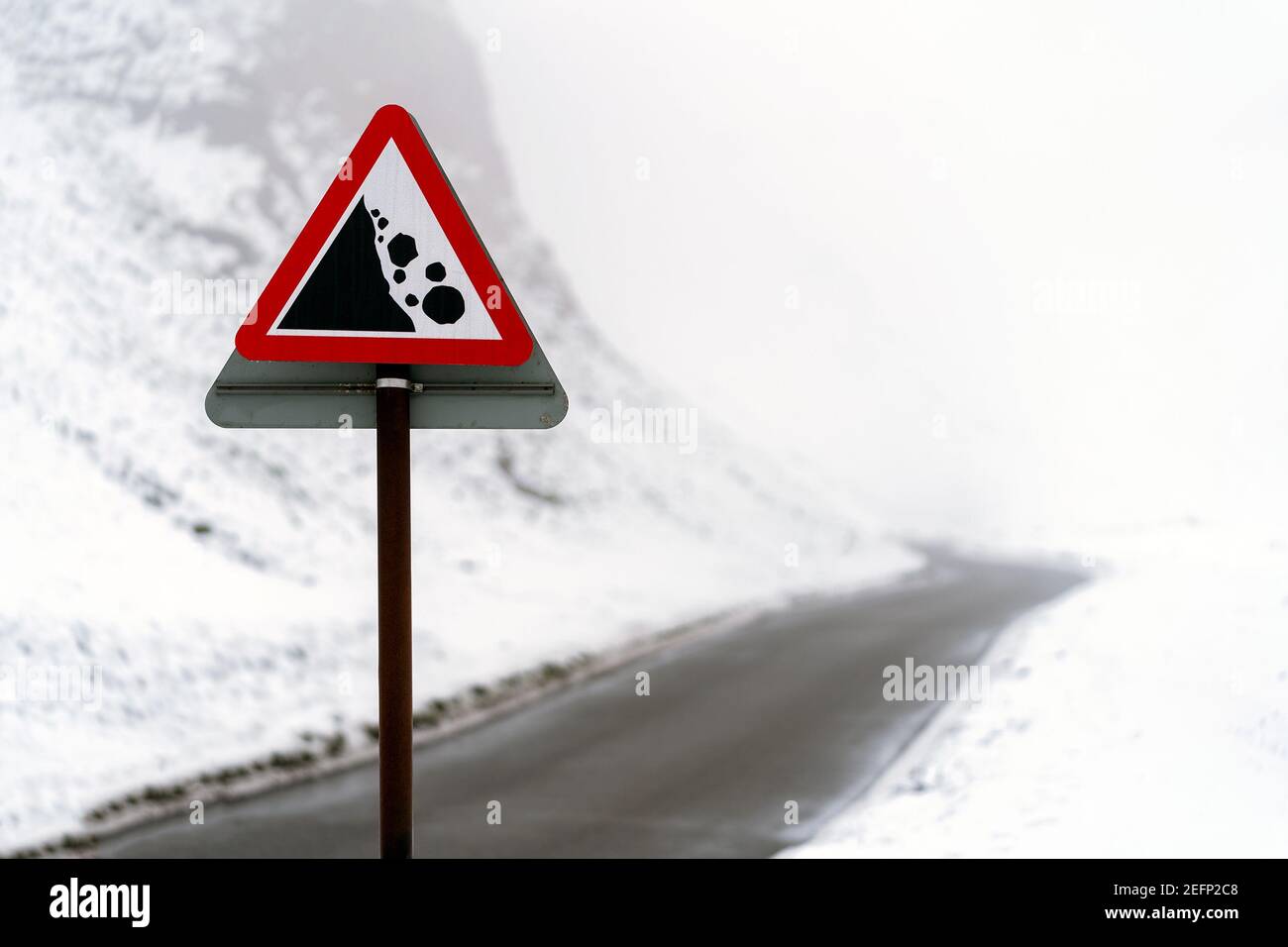 Panneau d'avertissement triangle rouge tombant sur de la neige glacée couverte route sinueuse de montagne passant par la brume brumeuse rempli de vallée entre collines en hiver Banque D'Images