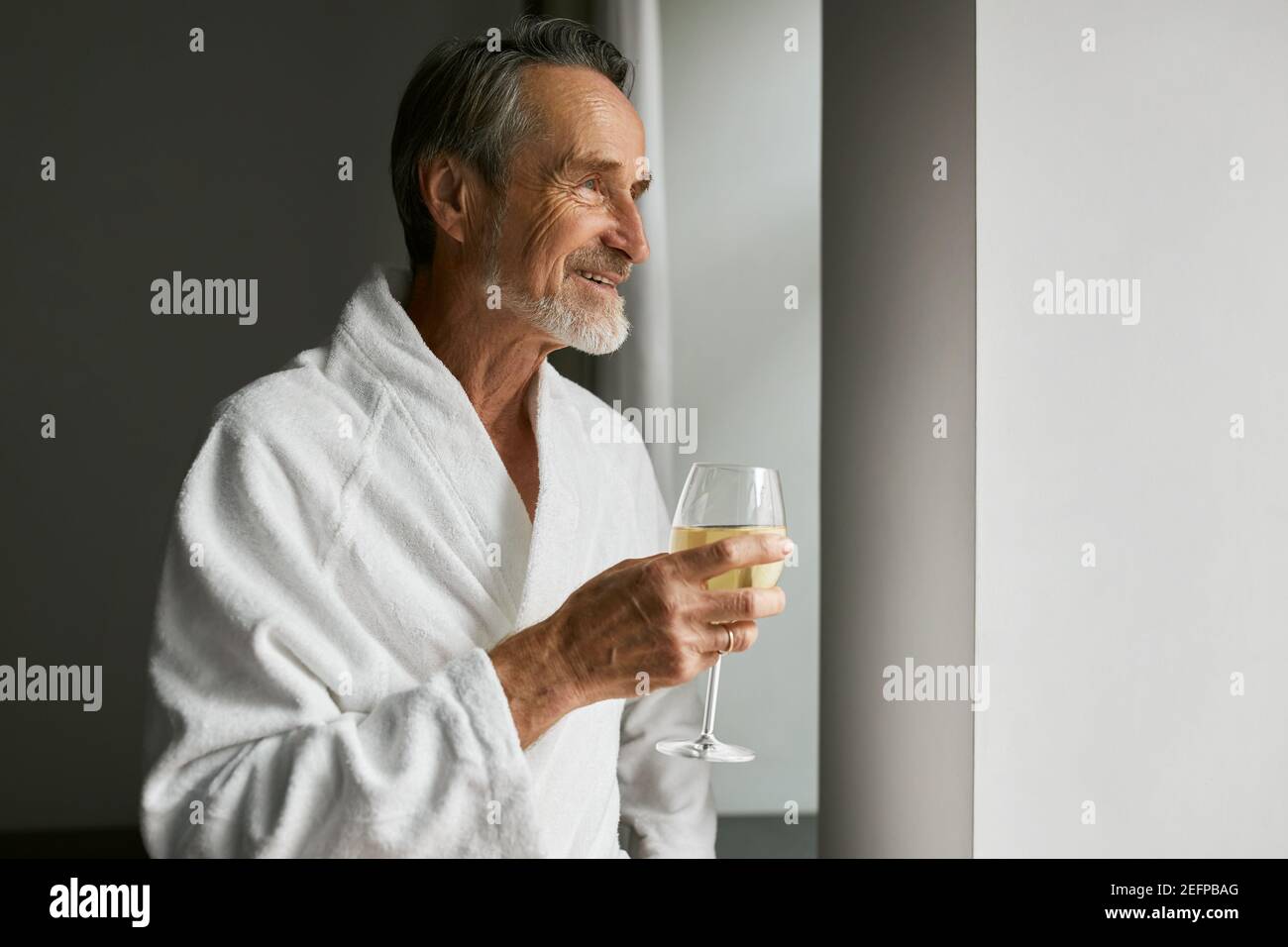 Homme senior souriant avec des lunettes de vin. Vue latérale d'un homme mature barbu portant un peignoir dans une chambre d'hôtel Banque D'Images