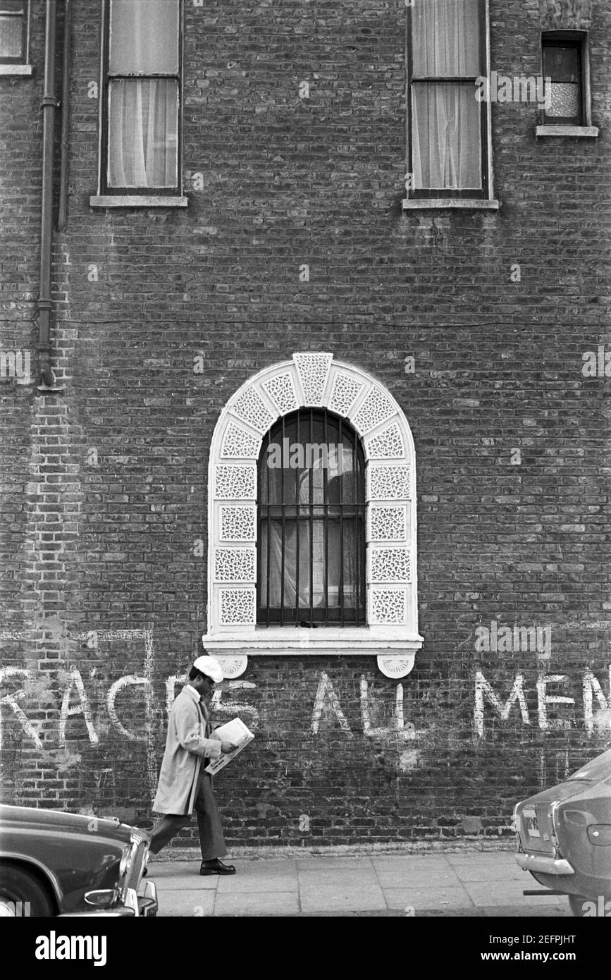 Royaume-Uni, West London, Notting Hill, 1973. Un homme noir lisant un journal passe devant un graffiti qui se lit comme «....races All Men.... ». Écrit sur les murs de Colville Houses, cul-de-sac, maintenant démoli et réconstruit. Banque D'Images