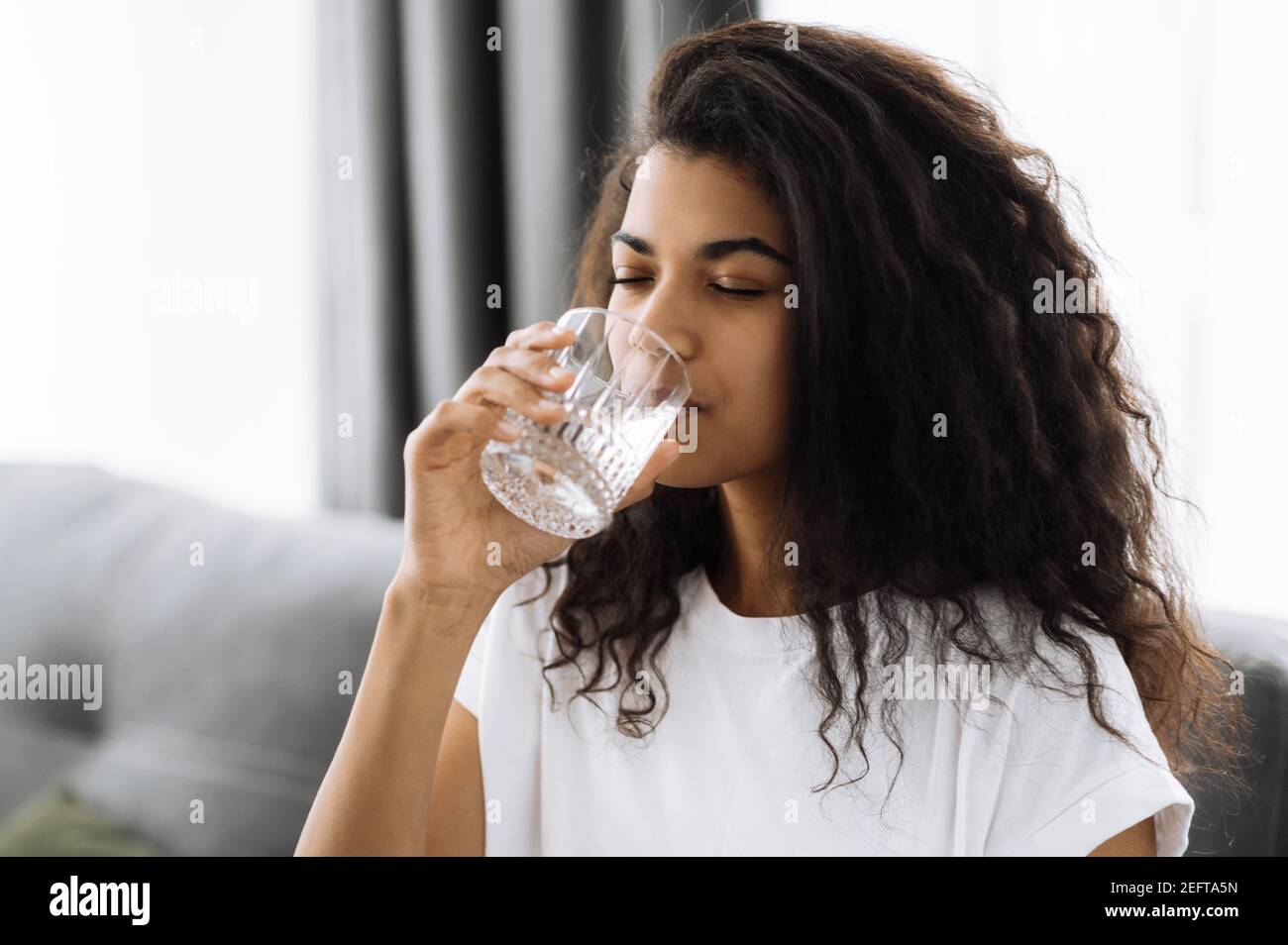 Un mode de vie sain. La jeune femme afro-américaine boit un verre d'eau tout en étant assise sur le canapé à la maison. Belle femme en bonne santé suivre un mode de vie sain Banque D'Images