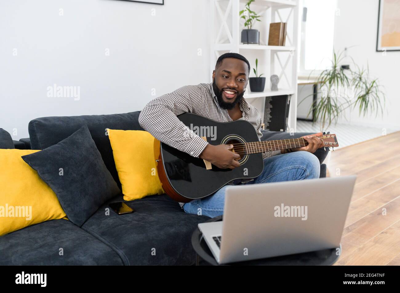 Souriant jeune Africain américain musicien de sexe masculin jouant de la guitare pendant un concert en ligne à la maison tout en étant isolé, donnant des cours virtuels, assis sur le Banque D'Images