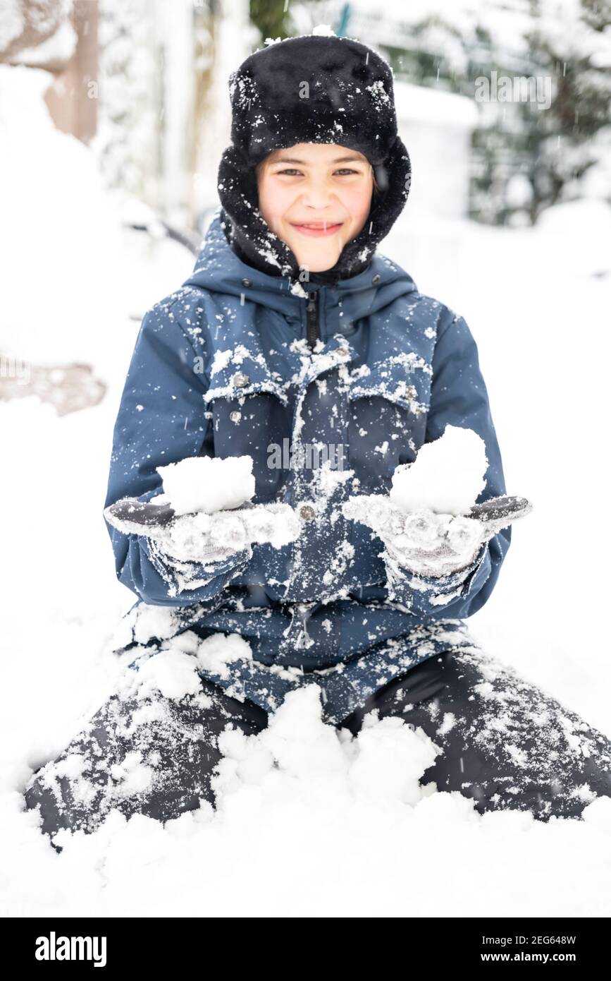 Mignon garçon dans les rabats chapeau assis dans la neige et faire des boules de neige. Adolescents s'amuser et profiter des vacances d'hiver. Mise au point sélective et virage Banque D'Images