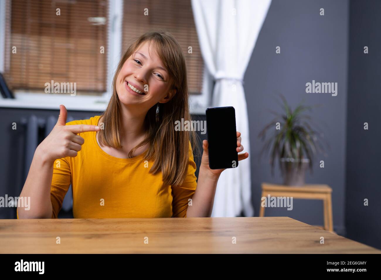 Femme blonde au bureau à domicile, souriante, tenant un smartphone moderne avec un écran maquette vierge, la femme adolescente pointe avec son index vers elle. Vide Banque D'Images