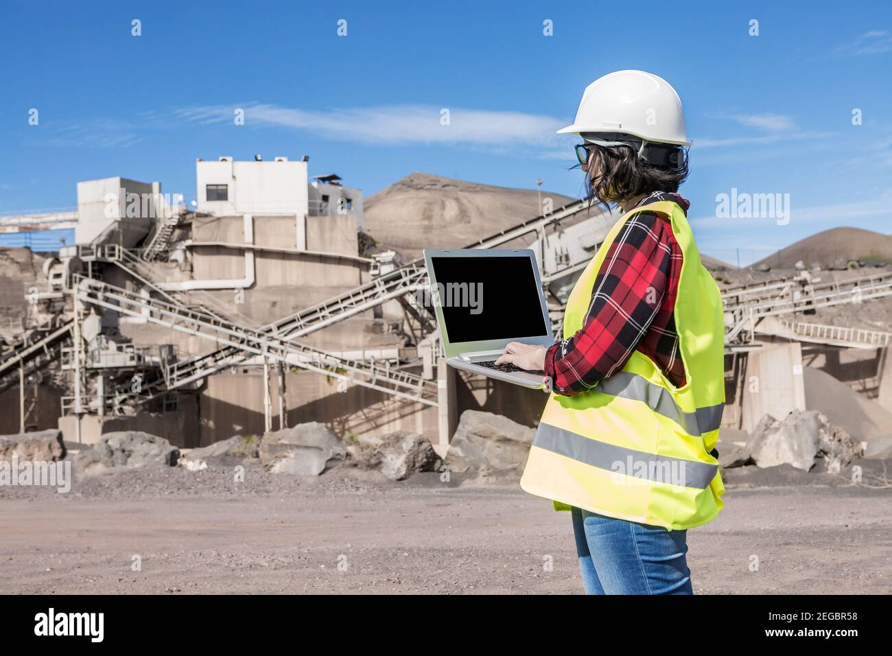 Vue latérale d'une femme ingénieur travaillant avec un casque et un gilet sur ordinateur portable avec écran noir lors de la visite du chantier de fa industriel non fini Banque D'Images
