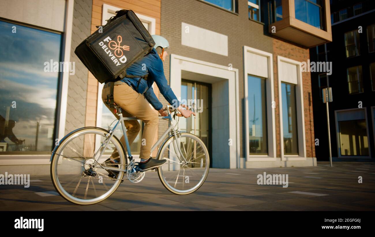 Happy Food Delivery Courier portant un sac à dos thermique fait un vélo sur la route pour livrer les commandes et les colis aux clients. Journée ensoleillée en ville avec Banque D'Images