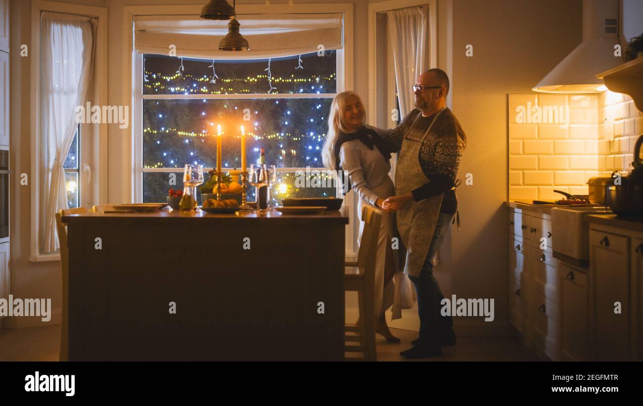 Heureux couple Senior en amour ont une soirée romantique, dansant dans la cuisine, célébrant l'anniversaire. Soirée romantique avec vin, table festive à l'intérieur Banque D'Images