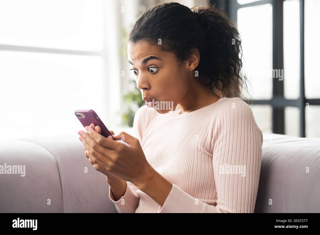 Surpris une adolescente afro-américaine assise sur le canapé, regardant le téléphone mobile, vérifiant les messages des médias sociaux ou naviguant sur Internet et Banque D'Images