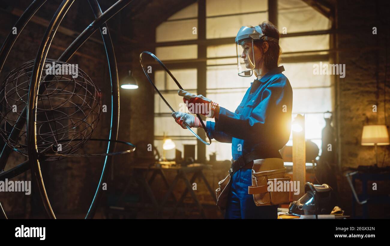Jeune artiste féminine contemporaine en combinaison bleue et masque de sécurité est en ajustement d'un anneau métallique à une sculpture de tube dans un atelier de studio. Femme habilitant Banque D'Images