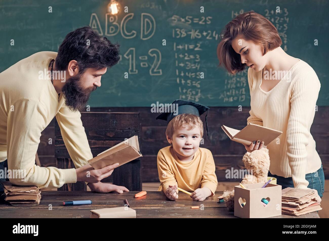 La famille se soucie de l'éducation de son fils. Père et mère lisant des livres, enseignant leur fils, tableau noir sur fond. Garçon enfant en tête de troisième cycle Banque D'Images