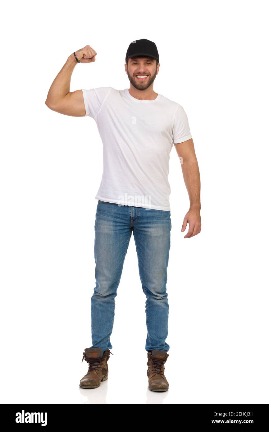 Décontracté jeune homme en casquette noire, chemise blanche, jeans et bottes brunes est debout, flexible biceps, regardant l'appareil photo et sourire. Vue avant. Longueur totale Banque D'Images
