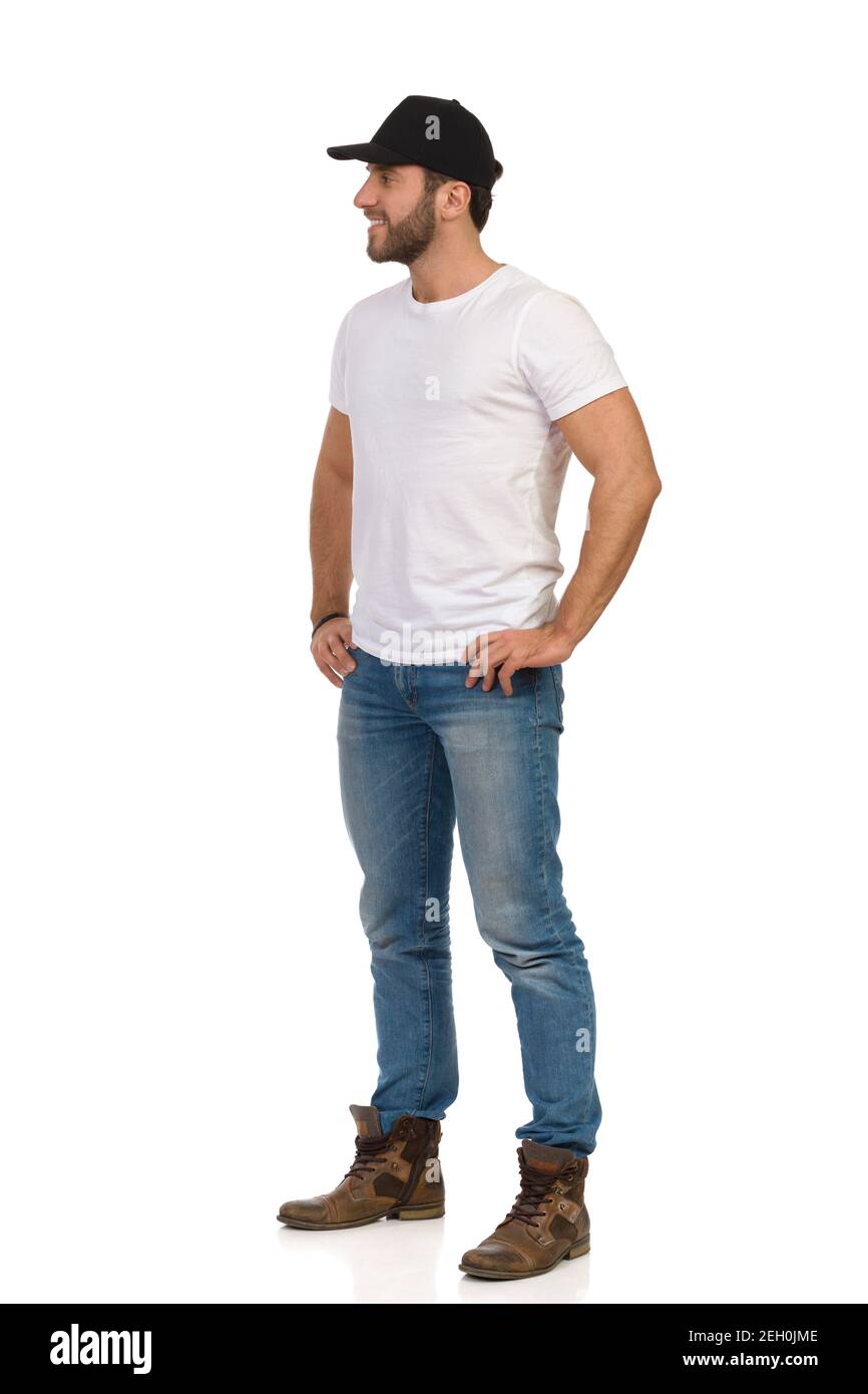 Décontracté jeune homme en casquette noire, chemise blanche, jeans et  bottes est debout avec les mains sur la hanche, regardant loin et souriant.  Prise de vue en studio pleine longueur isolée Photo