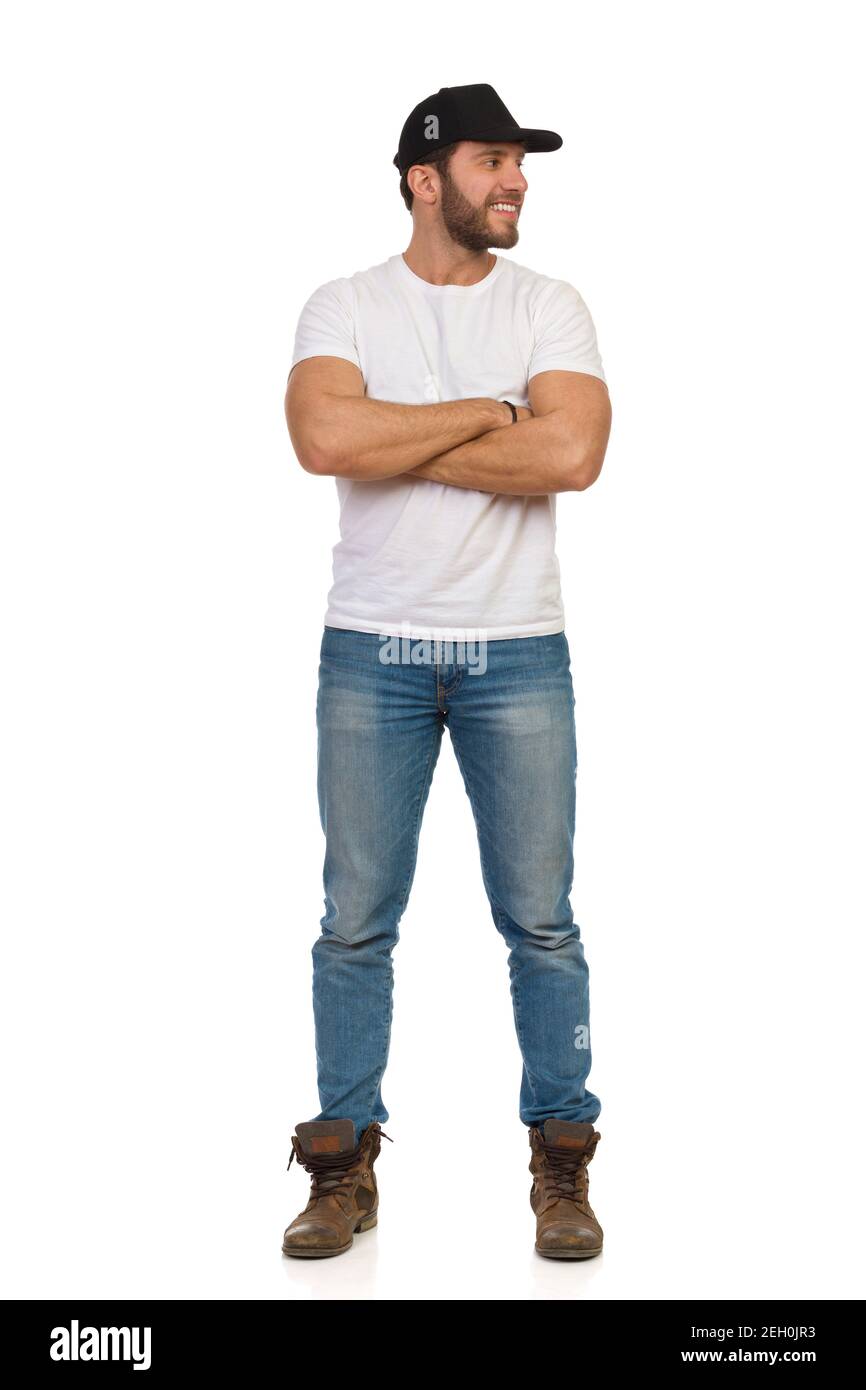 Décontracté jeune homme en casquette noire, jeans et t-shirt blanc est  debout avec les bras croisés, regardant le côté et souriant. Isolation de  la prise de vue en studio pleine longueur Photo