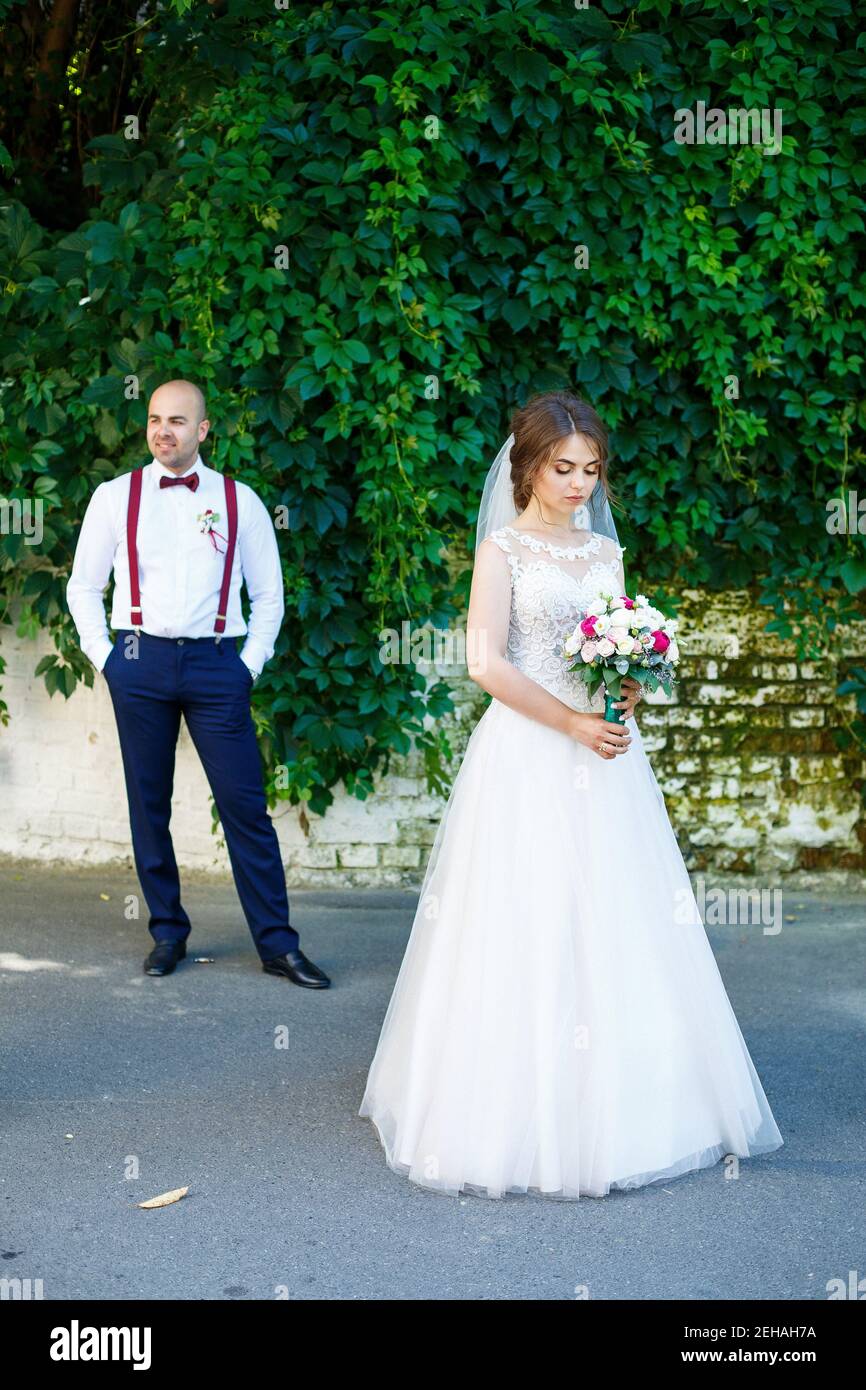 La mariée dans une robe blanche au premier plan et le marié avec bretelles  se tient derrière. Sur le mur d'arrière-plan avec des feuilles vertes Photo  Stock - Alamy