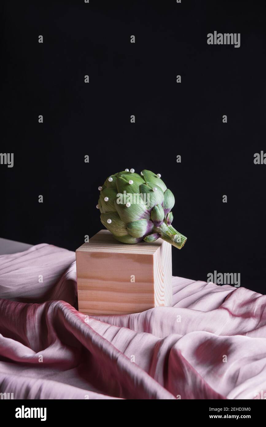Artichaut mûr placé sur un cube en bois sur une table avec de la soie tissu sur fond noir Banque D'Images