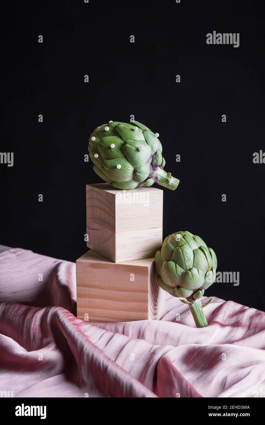 Artichauts mûrs placés sur un cube en bois sur une table avec de la soie tissu sur fond noir Banque D'Images