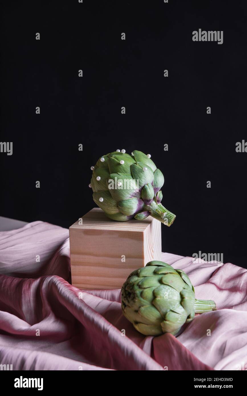 Artichauts mûrs placés sur un cube en bois sur une table avec de la soie tissu sur fond noir Banque D'Images