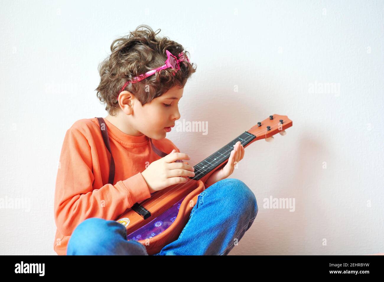 Drôle petit garçon avec des cheveux bouclés dans des verres en forme d'étoile rose joue la guitare jouet. Une star du rock joyeuse, hippie Banque D'Images
