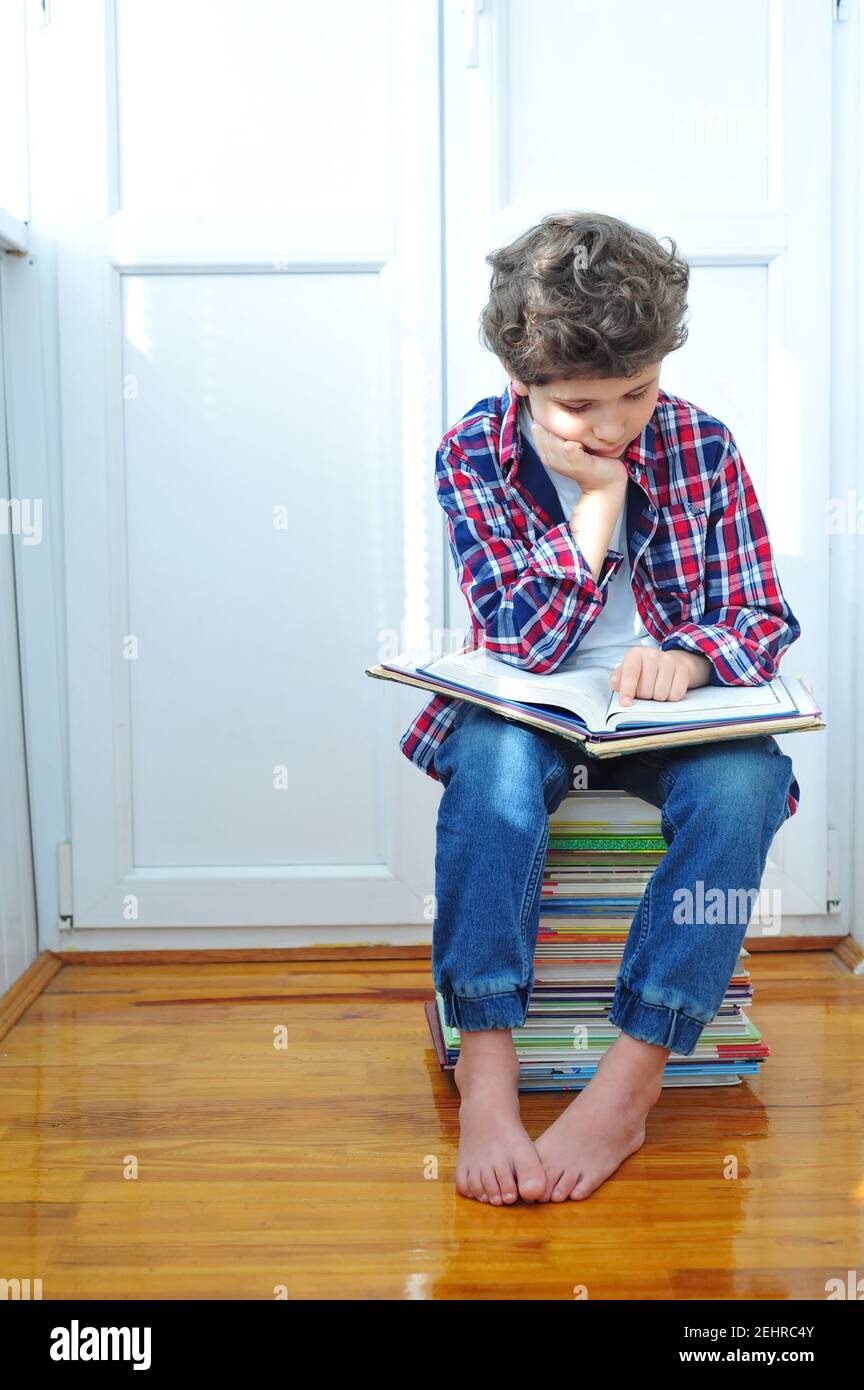 Un petit garçon mignon est assis dans sa chambre sur une pile de livres. Il était fatigué de lire. L'enfant a beaucoup de devoirs à faire. Banque D'Images