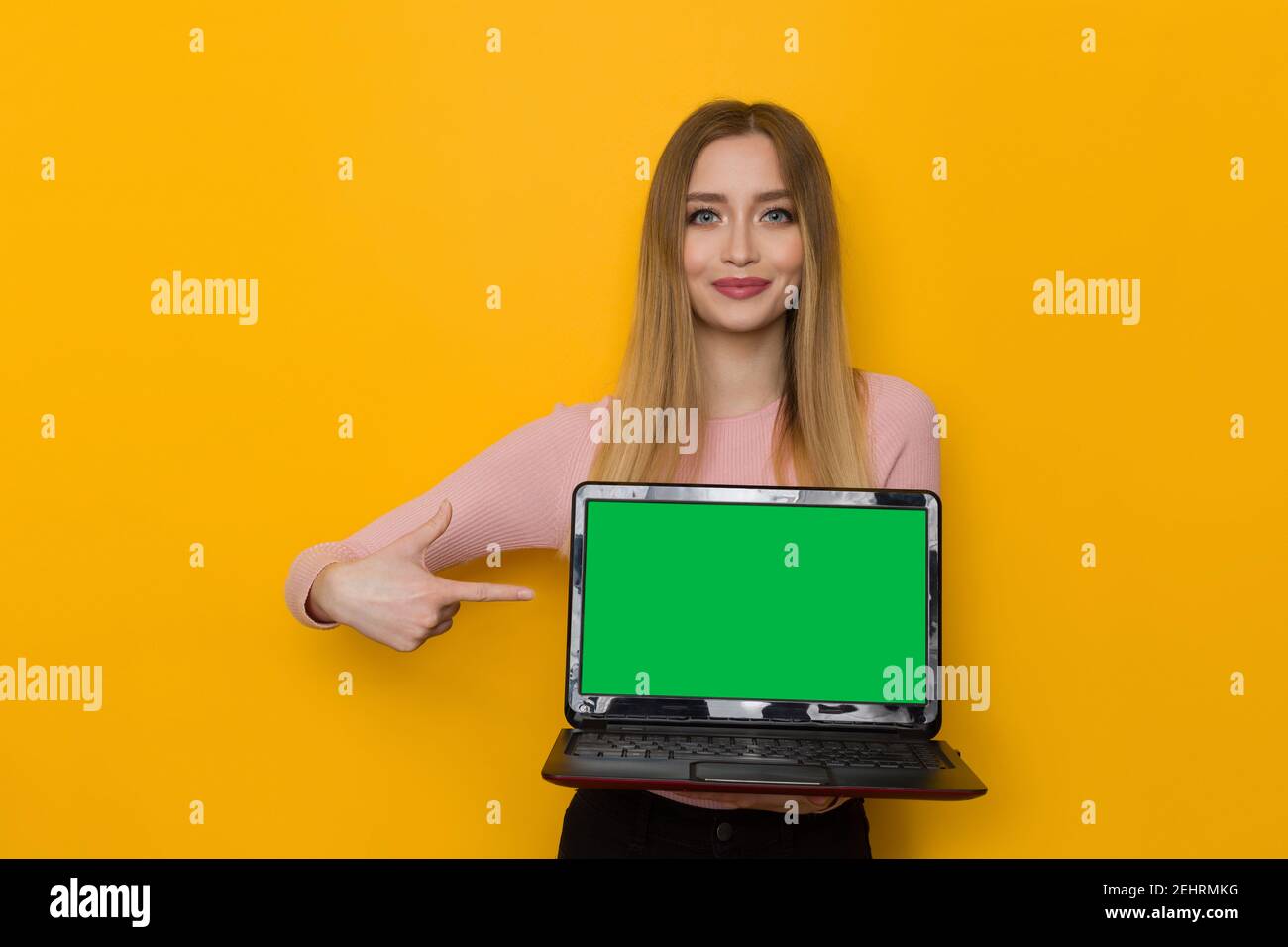 Une jeune femme souriante en chandail rose tient un ordinateur portable avec écran vert et le pointe vers lui. Taille haute studio tourné sur fond jaune. Banque D'Images