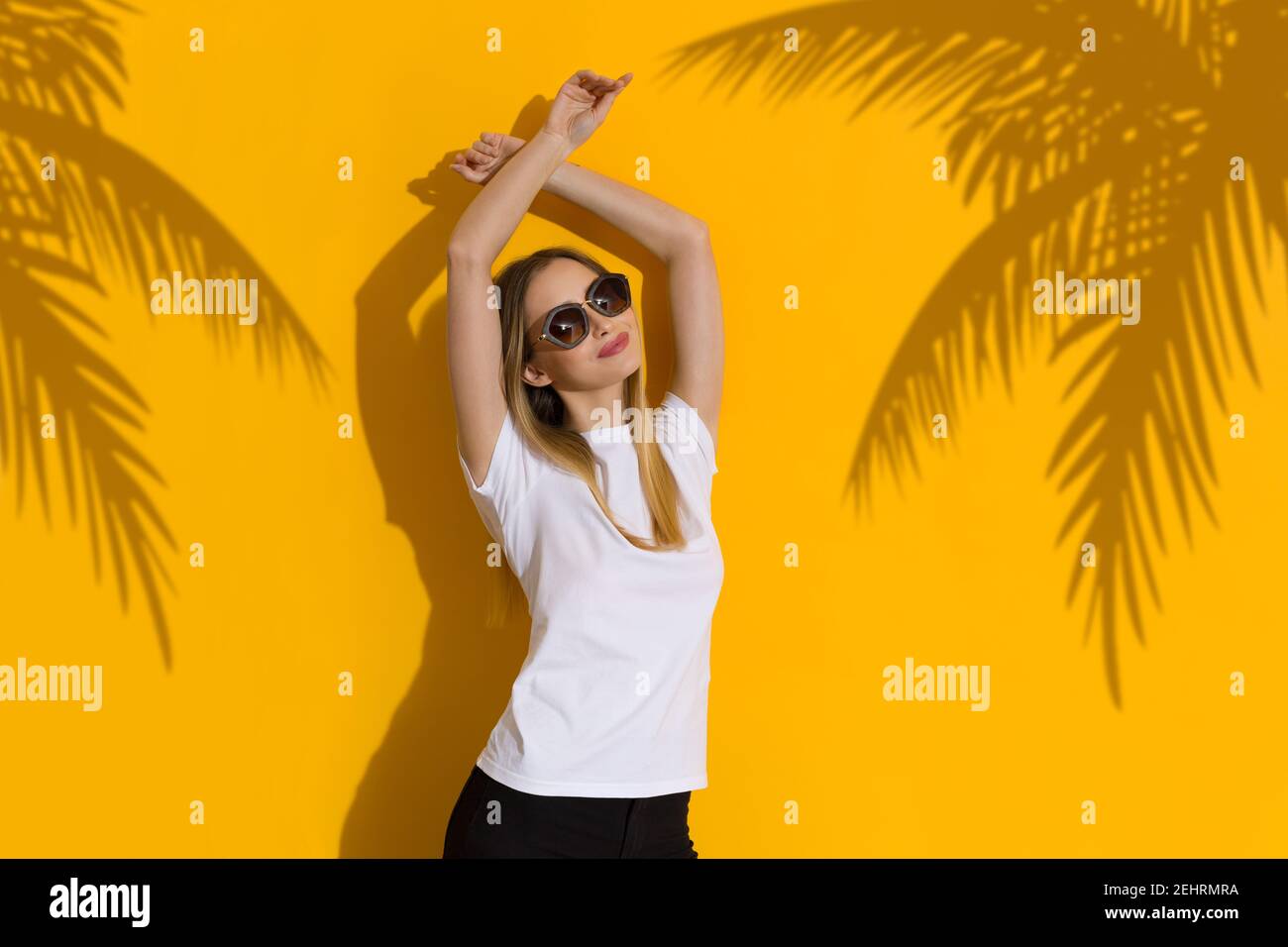 Bonne jeune femme blonde en chemise blanche et lunettes de soleil est debout avec les bras levés contre le mur jaune ensoleillé avec ombre des palmiers. Taille haute. Banque D'Images