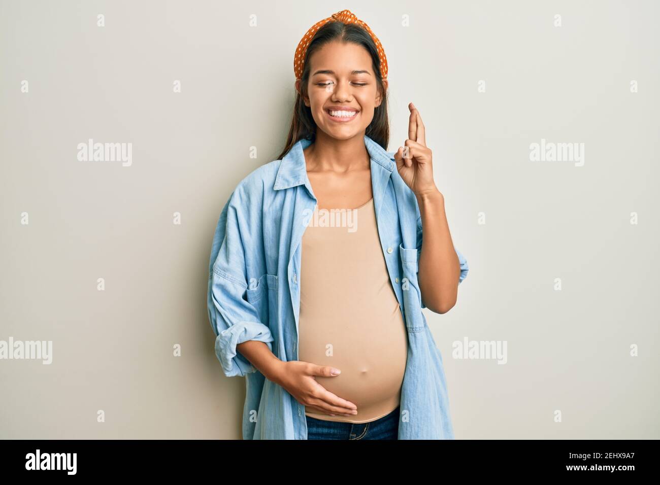 Belle femme hispanique s'attendant à un bébé, touchant le ventre enceinte gestante doigt croisé souriant avec espoir et les yeux fermés. Chance et superstitititieux Banque D'Images