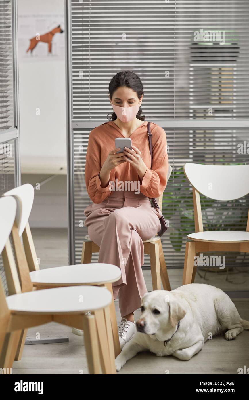 Portrait vertical de la jeune femme moderne avec chien en attente à la clinique vétérinaire et en utilisant un smartphone Banque D'Images