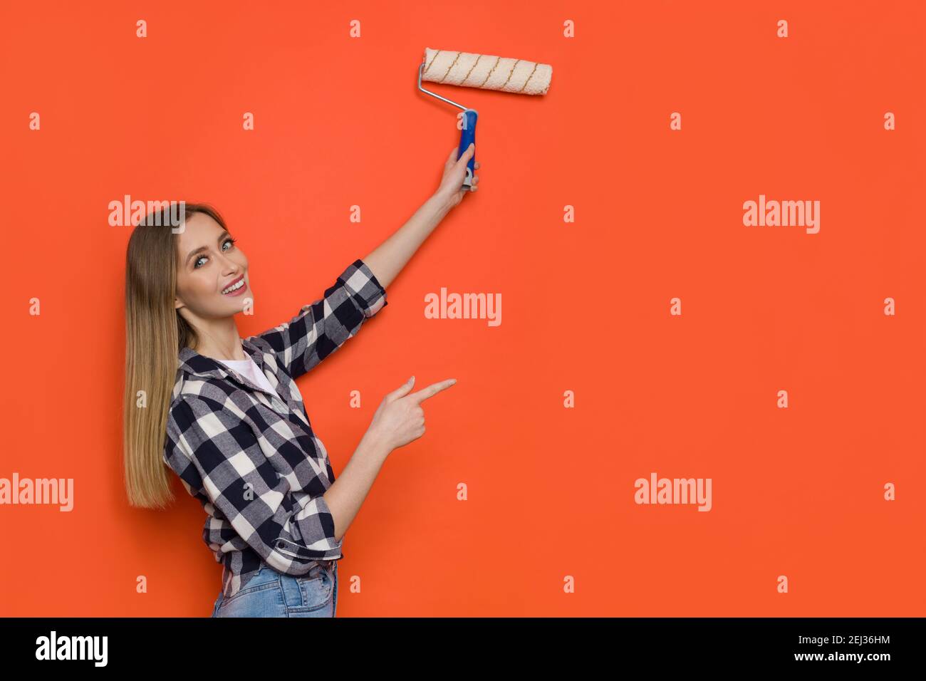 Jeune femme blonde en chemise de bûcherons et jeans peint le mur orange avec le rouleau de peinture, regardant la caméra sur l'épaule et pointant. Taille Banque D'Images