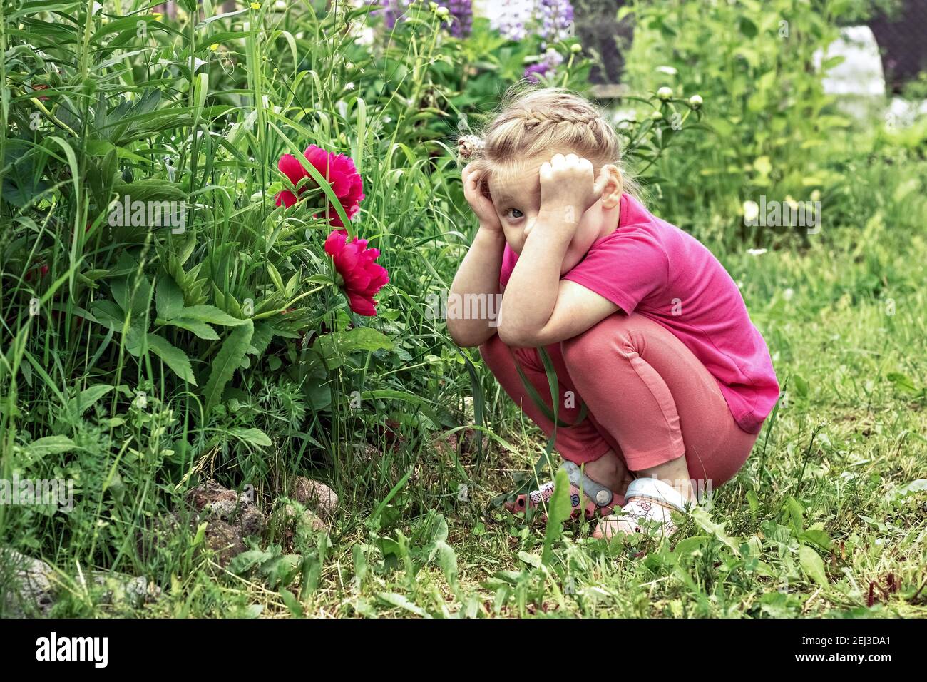 Une petite fille tenant sa tête en pensée, assise par un lit de fleur dans le jardin de pivoines roses. Banque D'Images