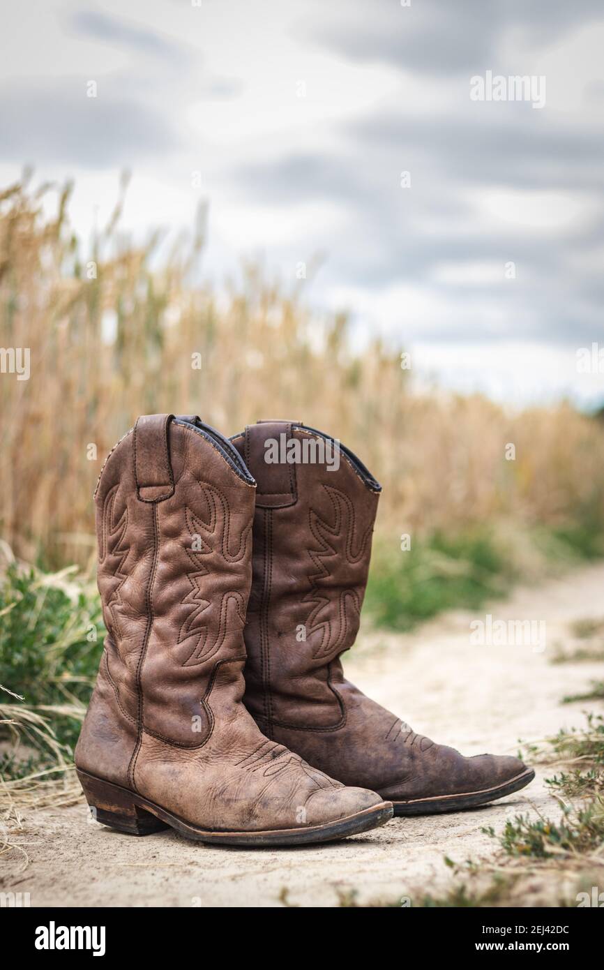Bottes de cowboy sur la route de terre à côté du champ de blé. Ancienne  chaussure en cuir usée à l'extérieur. Style rétro encore vie de chaussures  sur la scène rurale avec