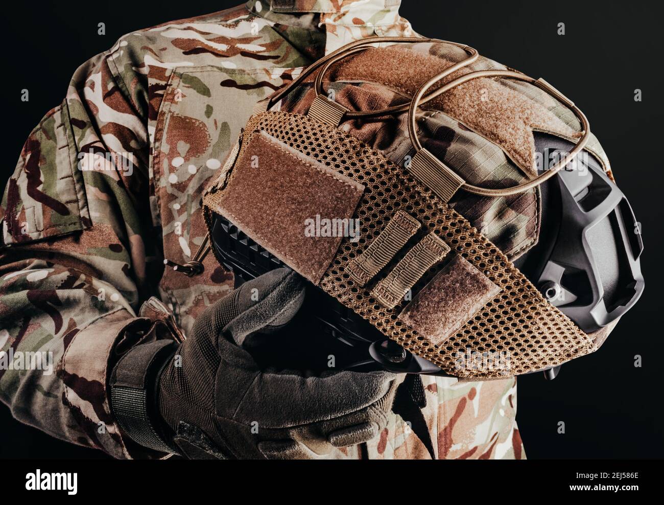 Photo d'un soldat en uniforme de camouflage et gants tactiques tenant un casque militaire avec une couverture sur fond noir. Banque D'Images