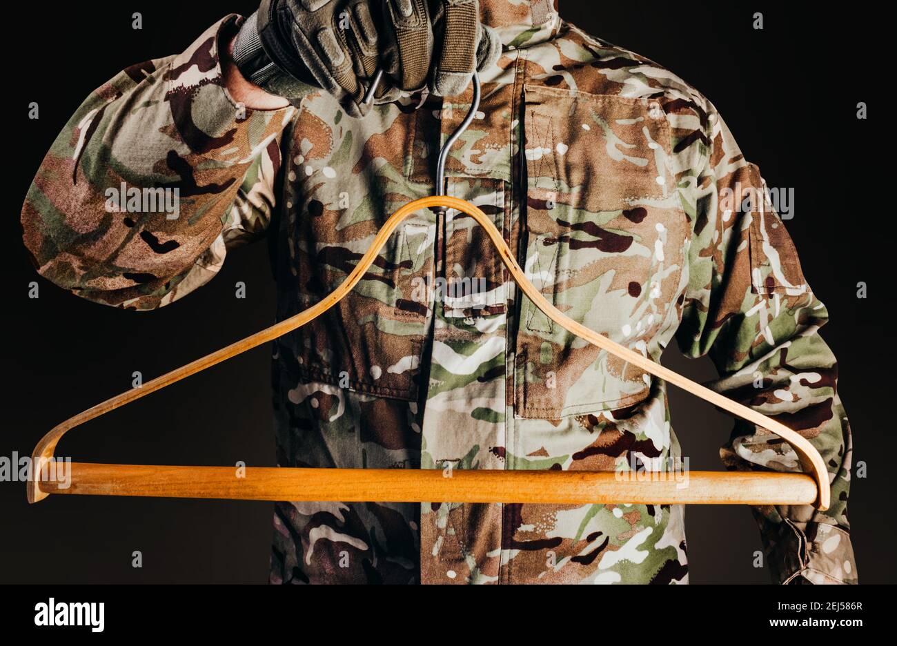 Photo d'un soldat en uniforme de camouflage et gants tactiques tenant un cintre de chemise en bois sur fond noir. Banque D'Images
