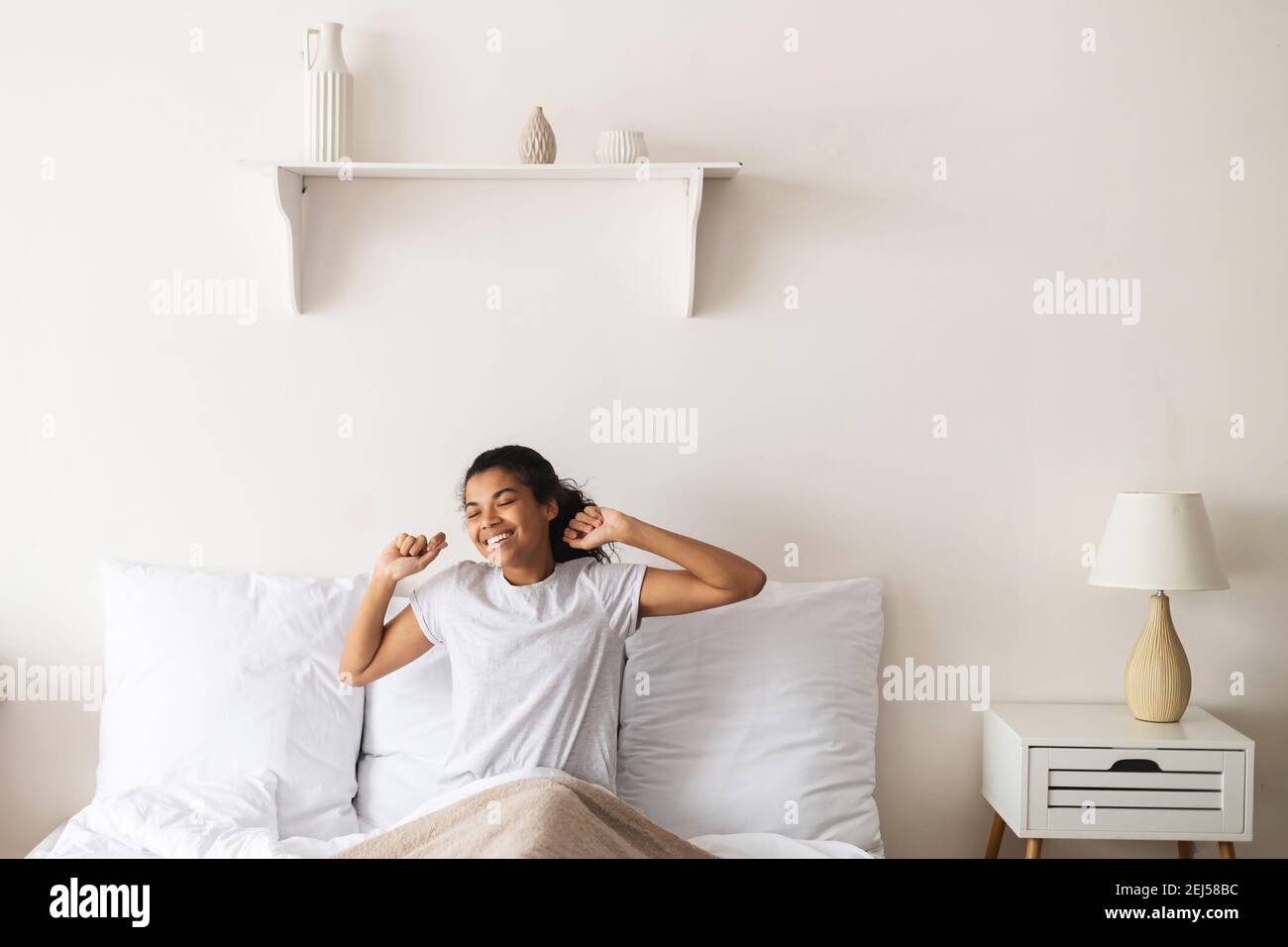 Joyeuse belle femme biraciale avec des cheveux sombres attachée assis dans le lit souriant, étirant, portant un t-shirt blanc simple, week-end, confortable et moderne Banque D'Images