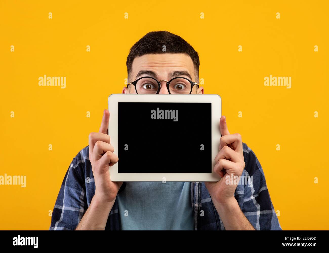 Un jeune homme choqué se cache derrière une tablette avec écran vide sur fond orange studio, maquette pour le design Banque D'Images