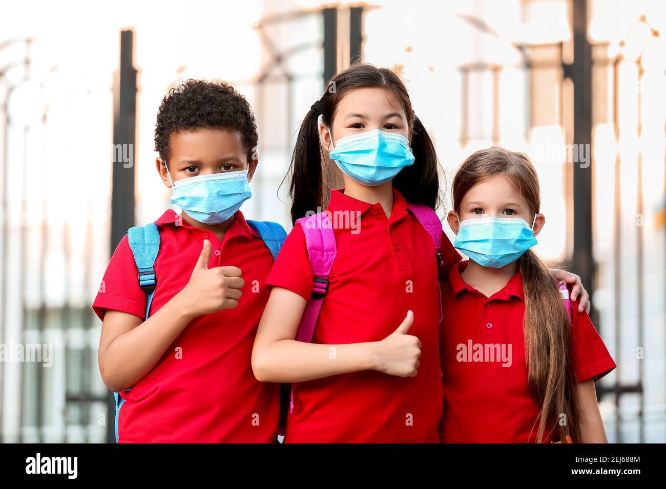 Petits élèves dans des masques médicaux montrant le pouce vers le haut près de l'école. Épidémie de coronavirus Banque D'Images