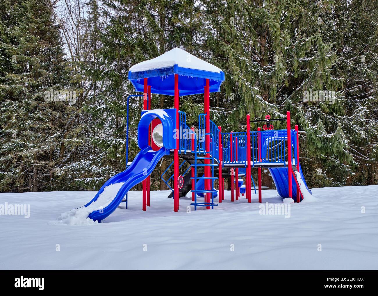 Le rouge vif et le bleu d'une structure de jeu pour enfants contraste fortement avec la neige blanche sur le sol et les arbres. Banque D'Images