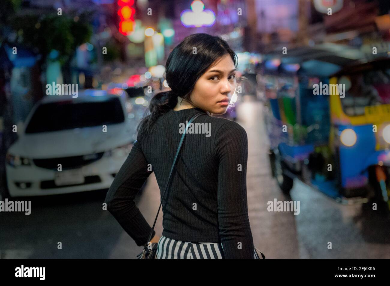 Jeune magnifique Asiangirl dans une rue animée la nuit, marchant tout en regardant de nouveau la caméra. Lumières de la ville et tuk-tuk en arrière-plan. Banque D'Images