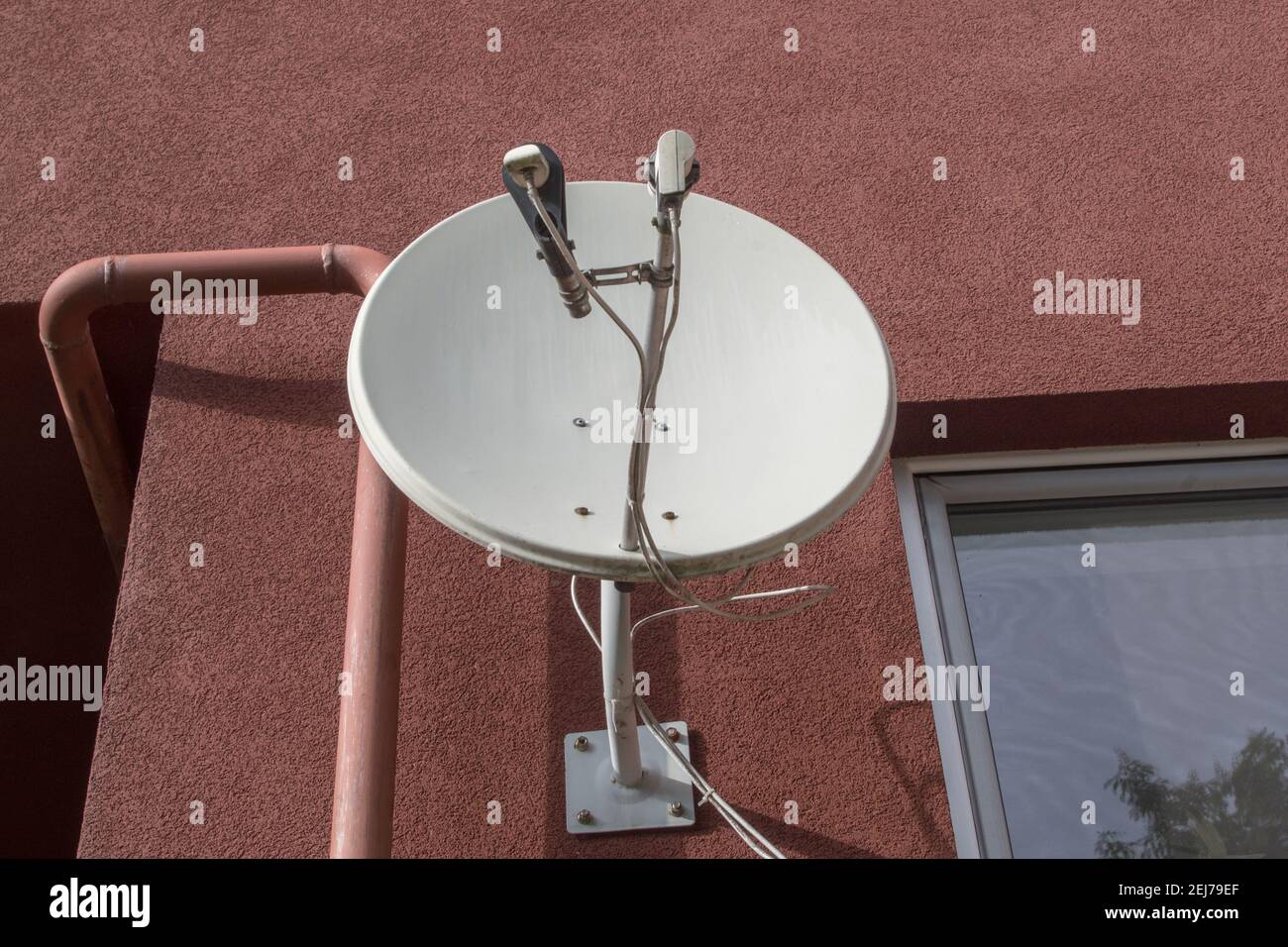 Antenne de télévision par satellite ronde en métal blanc fixée par des boulons d'ancrage à la façade marron d'un immeuble à gauche de la fenêtre et à droite de Banque D'Images