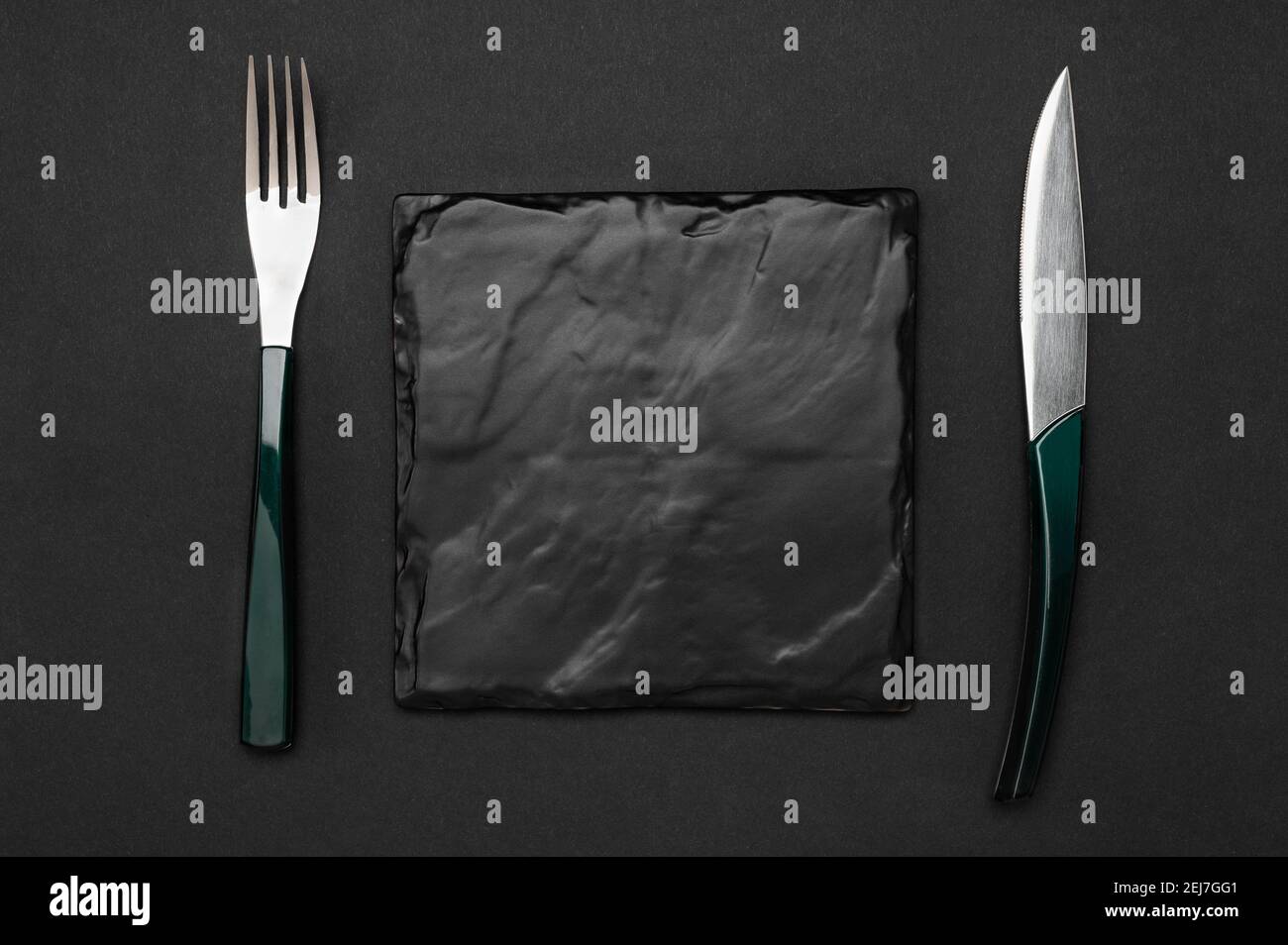 Couteau et fourchette avec plaque carrée en ardoise noire pour le texte du menu ou de la recette sur une table noire. Maquette un tableau vierge et des couverts sur fond de table noir Banque D'Images
