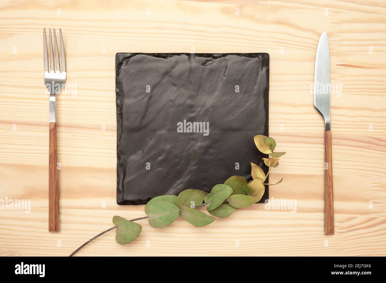 Couteau et fourchette avec plaque carrée en ardoise noire pour le texte du menu ou de la recette et branche d'eucalyptus d'or sur table en bois. Maquette planche vierge et couverts sur la lune Banque D'Images