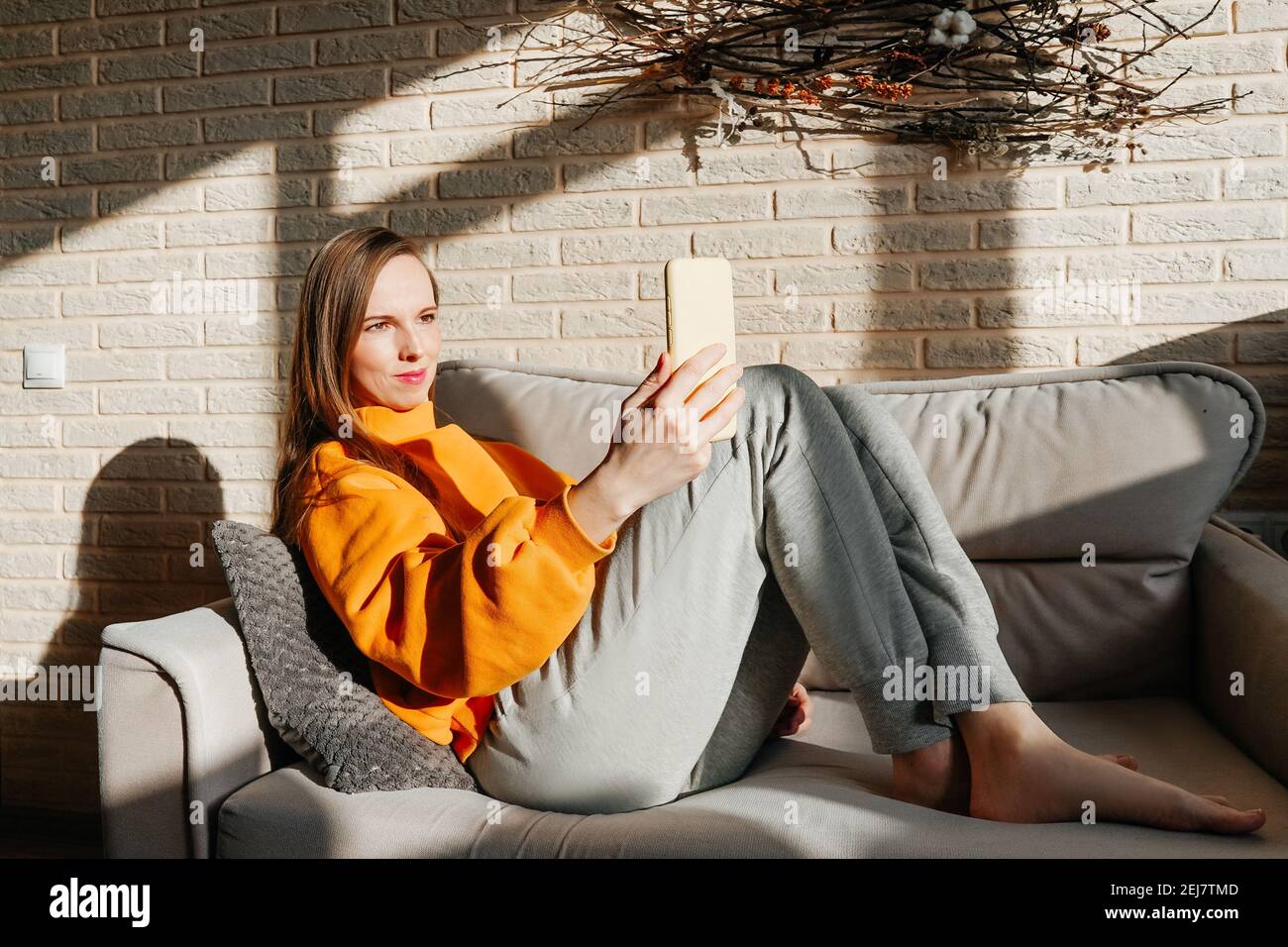 Jolie jeune femme avec un selfie assis sur un canapé à la maison. Sweat-shirt  orange jaune vif et pantalon gris. Mur de briques et pannel naturel sur le  panneau arrière Photo Stock -