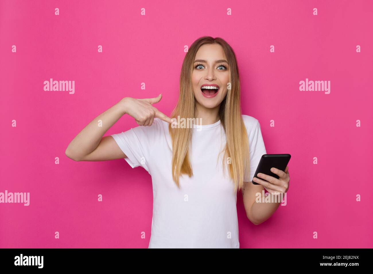 Une jeune femme heureuse en chemise blanche tient le téléphone, le pointe vers lui et crie. Taille haute studio tourné sur fond rose. Banque D'Images