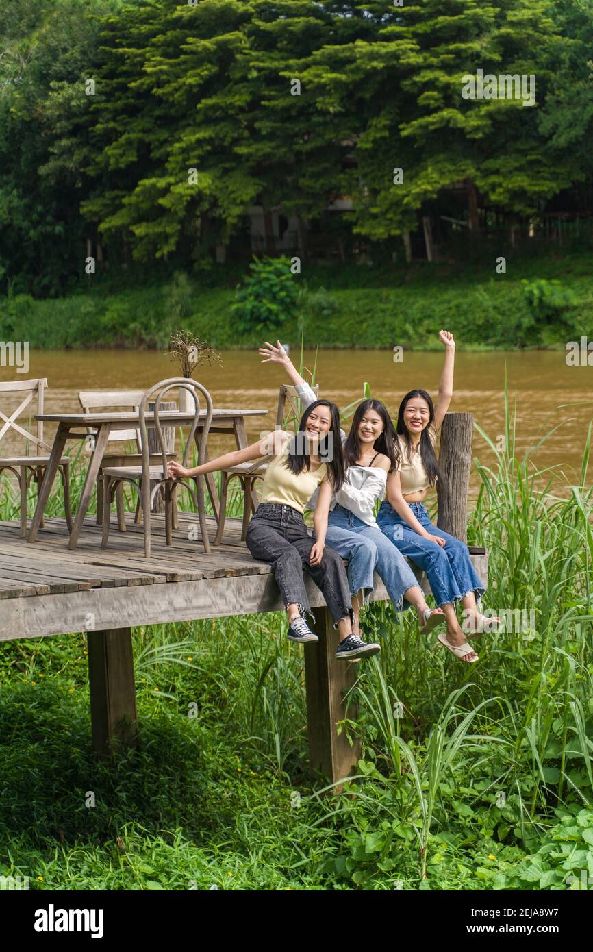Trois jeunes amies asiatiques heureuses s'asseyant sur le pont en bois au bord d'une rivière avec de grands arbres en arrière-plan, amitié ou jeune ha Banque D'Images