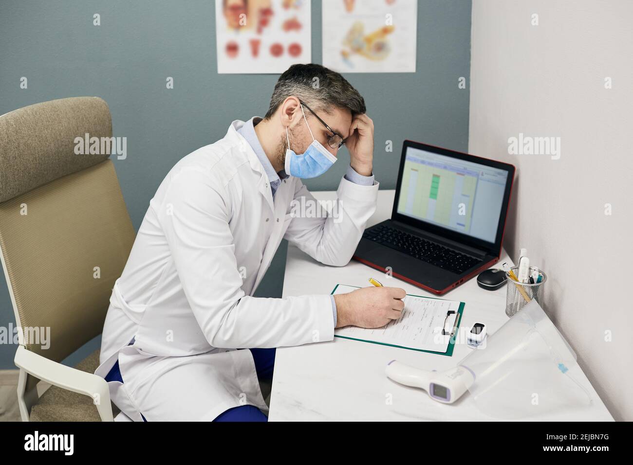 Un médecin de sexe masculin sérieux remplit un formulaire de dossier médical sur son lieu de travail dans une clinique médicale. Médecin portant un manteau blanc et un masque de protection Banque D'Images